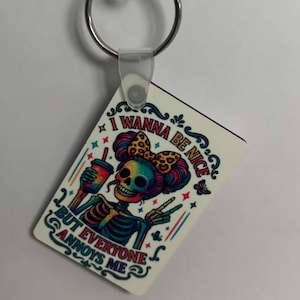 May include: A rectangular keychain with a colorful illustration of a skeleton with a bow, holding a drink, and making a peace sign. The text on the keychain reads "I WANNA BE NICE BUT EVERYONE ANNOYS ME."
