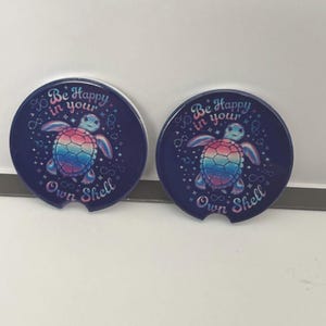 Puede incluir: Dos posavasos para coche azul marino con un diseño de tortuga marina colorida. Los posavasos tienen el texto "Be Happy in your Own Shell" en una fuente caprichosa. El caparazón de la tortuga es un degradado arcoíris.