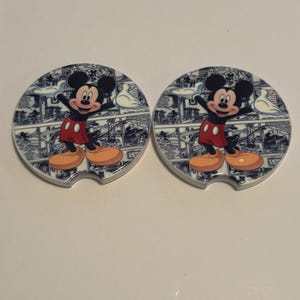 May include: Two round car coasters featuring Mickey Mouse in a classic pose. The coasters have a blue and white patterned background with a cartoon illustration of Mickey Mouse in red shorts, yellow shoes, and black ears.