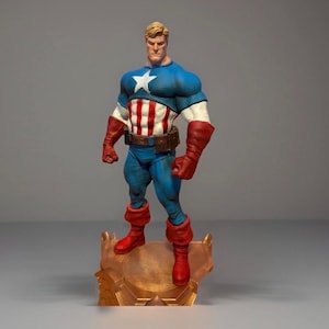 Pu&ograve; includere: Una statuetta di Captain America su una base color oro. La figura indossa un costume blu e bianco con una stella bianca sul petto, strisce rosse e bianche e guanti e stivali rossi. La statuetta ha i capelli biondi e un'espressione determinata.