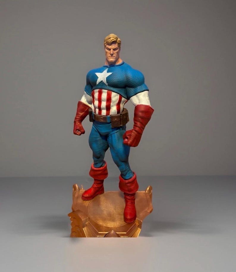 Pu&ograve; includere: Una statuetta di Captain America su una base color oro. La figura indossa un costume blu e bianco con una stella bianca sul petto, strisce rosse e bianche e guanti e stivali rossi. La statuetta ha i capelli biondi e un'espressione determinata.