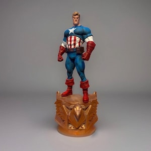 Pu&ograve; includere: Una statuetta di Captain America su una base decorativa. La figura indossa un costume blu, bianco e rosso con una stella sul petto. La base &egrave; dorata e presenta un design ornato.