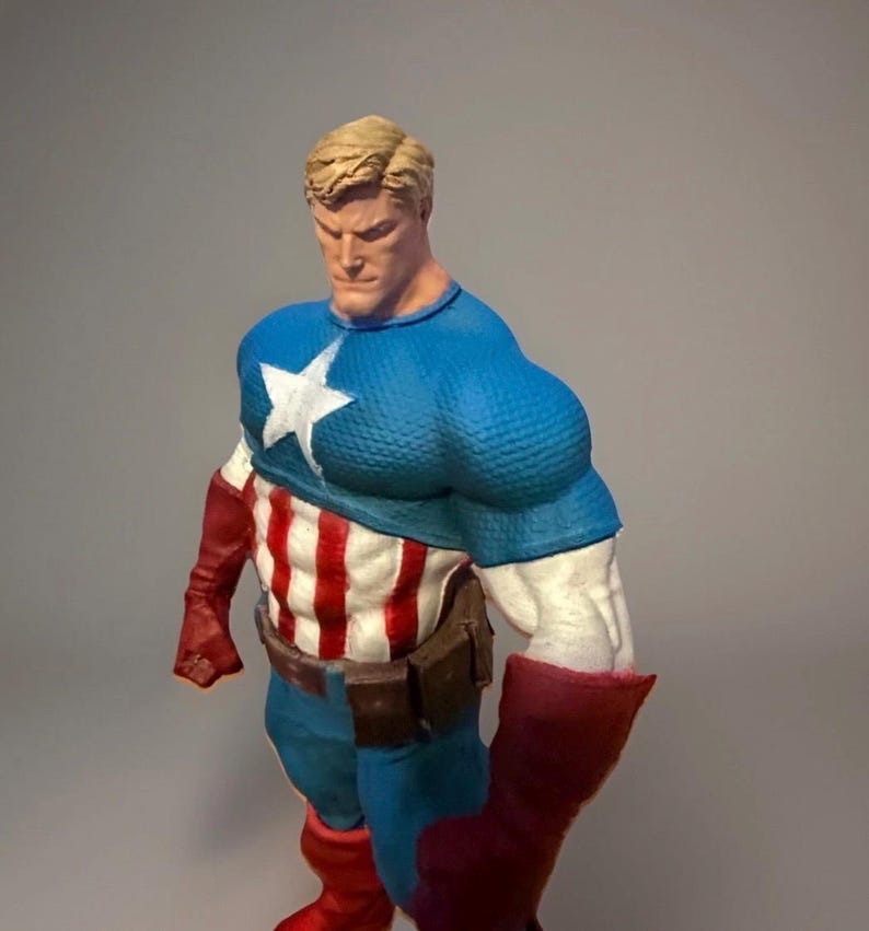 Pu&ograve; includere: Una statuina di Captain America in posa classica. La figura indossa un top blu con una stella bianca, un busto a strisce rosse e bianche e stivali rossi. La figura ha i capelli biondi e un'espressione determinata.
