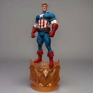 Pu&ograve; includere: Una statuetta di Captain America su una base dorata. La figura indossa un top blu e bianco con una stella, un busto a righe rosse e bianche, pantaloni blu, stivali rossi e guanti marroni. La base ha un design ornato.