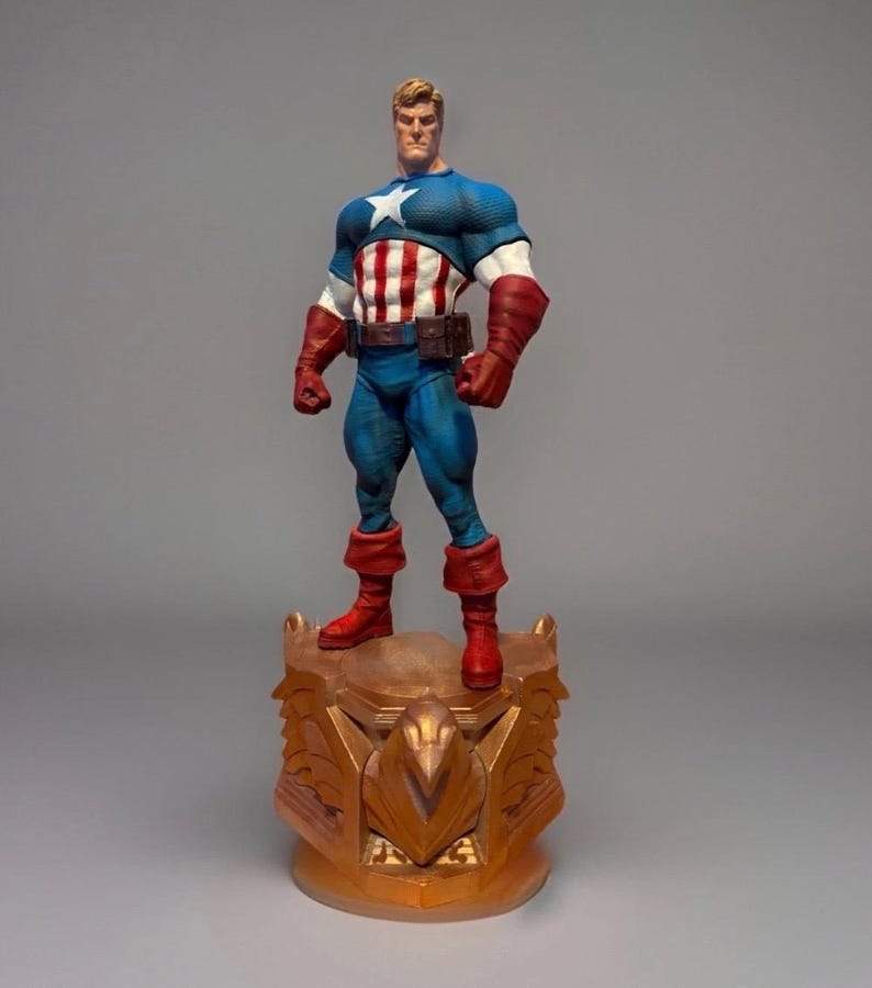 Pu&ograve; includere: Una statuetta di Captain America su una base dorata. La figura indossa un top blu e bianco con una stella, un busto a righe rosse e bianche, pantaloni blu, stivali rossi e guanti marroni. La base ha un design ornato.