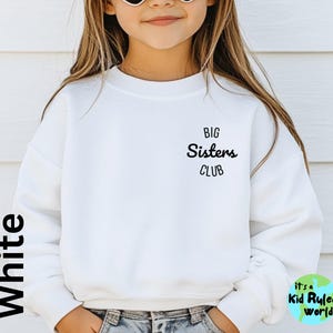 May include: White sweatshirt with the text "Big Sisters Club" in black lettering. The sweatshirt is being worn by a child.