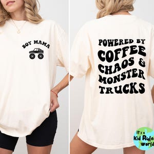 May include: A cream-coloured t-shirt featuring black text and graphics. The front displays "Boy Mama" above a monster truck illustration. The back reads "Powered by Coffee Chaos & Monster Trucks". Part of the "Kid Ruled World" collection.