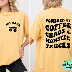 May include: Mustard yellow t-shirt with black text and graphics. The front reads "Boy Mama" above a monster truck graphic. The back says "Powered by Coffee Chaos & Monster Trucks". The shirt is a casual, oversized fit.