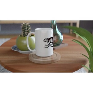 May include: A white ceramic mug with a black graphic of a rat and the text "Automotive". The mug is on a wooden coaster. A small cactus in a green pot and a decorative glass bottle are in the background.