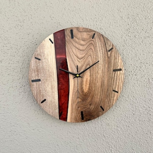 May include: A round wooden wall clock with a red resin inlay. The clock has black hands and black Roman numeral markings.