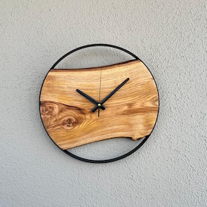 Puede incluir: Un reloj de pared redondo de madera con una esfera de veta de madera natural y manecillas negras. La esfera del reloj está encerrada en un anillo de metal negro. El reloj está montado en una pared gris texturizada.