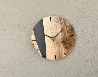 Puede incluir: Un reloj redondo de madera con detalles de resina epoxi negra. La esfera del reloj tiene agujas de hora y minuto negras y un centro dorado.