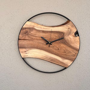 May include: A round wooden clock with a black metal frame. The clock face is made of a natural wood slice with a unique grain pattern. The clock hands are black.