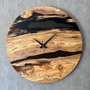 May include: Round wooden wall clock with a natural wood grain pattern and black epoxy resin accents. The clock features black hands and a simple, modern design. The clock's diameter is approximately 12 inches.