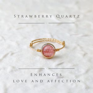 Op de afbeelding: Een gouden ring met een roze aardbeienkwarts edelsteen. De ring is omwikkeld met gouden draad en de tekst "STRAWBERRY QUARTZ" staat boven de ring. De tekst "ENHANCES LOVE AND AFFECTION" staat onder de ring.