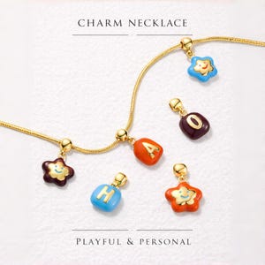 Puede incluir: Un collar con colgantes de color dorado con colgantes de esmalte de colores. Los colgantes incluyen letras y formas de estrella con caras sonrientes. También se ve el texto "CHARM NECKLACE" y "PLAYFUL & PERSONAL".
