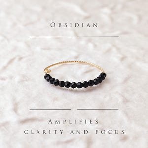 Könnte beinhalten: Ein goldener Ring mit schwarzen Obsidianperlen. Der Ring liegt auf einem weißen, strukturierten Hintergrund. Der Text "OBSIDIAN" befindet sich über dem Ring und der Text "AMPLIFIES CLARITY AND FOCUS" unter dem Ring.