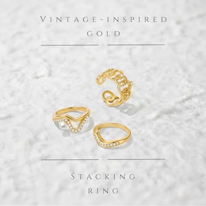 Peut inclure: Trois bagues empilables dorées d'inspiration vintage. L'une est ornée d'une perle, une autre a une forme en V avec de petits cristaux, et la troisième est de style chaîne avec des cristaux. Le texte "VINTAGE-INSPIRED GOLD" et "STACKING RING" est également visible.