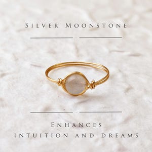 Puede incluir: Un anillo de piedra lunar plateada con un envoltorio de alambre dorado. El anillo está sobre un fondo blanco con el texto "SILVER MOONSTONE" y "ENHANCES INTUITION AND DREAMS".