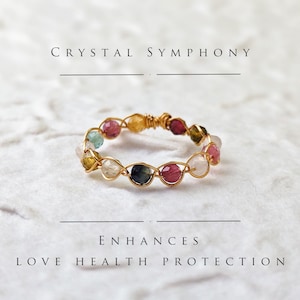 Puede incluir: Anillo envuelto en alambre dorado con una variedad de piedras preciosas de colores, incluyendo rosa, verde, azul y amarillo. El texto "CRYSTAL SYMPHONY" está en la parte superior de la imagen y "ENHANCES LOVE HEALTH PROTECTION" está en la parte inferior.