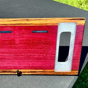 May include: A wooden key holder with a purple and brown wood grain finish. It has two hooks and three slots for keys. The slots are lined with a white material.