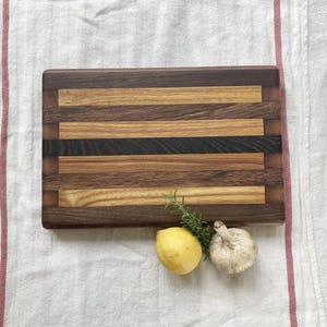 May include: A rectangular wooden cutting board with a striped pattern of light and dark wood. The board is made of walnut, maple, and cherry wood. It is sitting on a white linen tablecloth with a lemon and garlic clove beside it.