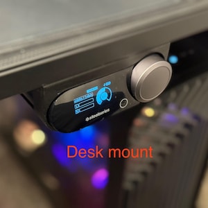 May include: Black desk mount with a digital display and a large control knob. The display shows blue illuminated graphics and text, with the SteelSeries logo. The mount is attached to a black surface.