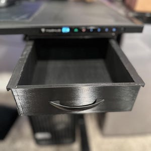 May include: A black drawer with a black handle is open and partially extended from a black desk. The drawer is empty.