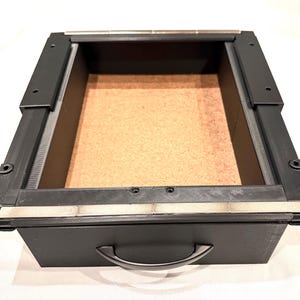 May include: A rectangular black storage drawer featuring a smooth matte finish, a curved handle on the front, and a light brown interior base. The drawer includes integrated black side rails with metallic silver accents along the top edges.