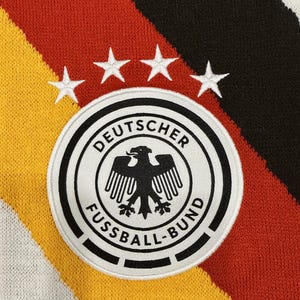Può includere: Un tessuto lavorato a maglia con strisce diagonali gialle, rosse e nere. Al centro, una toppa circolare bianca presenta un'aquila nera e il testo DEUTSCHER FUSSBALL-BUND, sormontato da quattro stelle bianche ricamate disposte ad arco.
