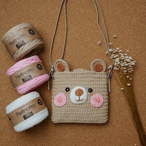 Peut inclure: Un sac bandouli&egrave;re crochet&eacute; en forme d'ours beige avec une longue sangle, dot&eacute; d'un museau blanc, de joues roses et d'oreilles marron. &Agrave; c&ocirc;t&eacute; se trouvent trois bobines de fil Fischer Refinatto beige, rose et blanc, et des fleurs s&eacute;ch&eacute;es.