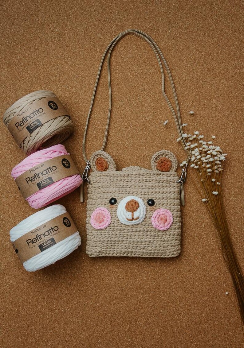 Peut inclure: Un sac bandouli&egrave;re crochet&eacute; en forme d'ours beige avec une longue sangle, dot&eacute; d'un museau blanc, de joues roses et d'oreilles marron. &Agrave; c&ocirc;t&eacute; se trouvent trois bobines de fil Fischer Refinatto beige, rose et blanc, et des fleurs s&eacute;ch&eacute;es.