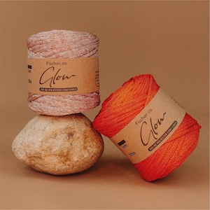 May include: Two spools of yarn, one orange and one peach, with brown paper labels that read "Fischer.co Glow". The yarn is displayed on a light brown rock against a brown background. The text "FIO DE POLIESTER COM LUREX" is also visible.
