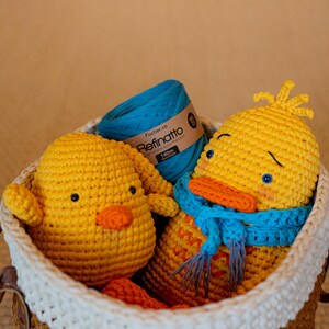 Peut inclure: Deux canetons au crochet jaunes, dont l'un porte une &eacute;charpe bleue, et une pelote de fil bleu sarcelle avec une &eacute;tiquette Fischer.co Refinatto 140m, le tout dans un panier tress&eacute; avec un bord blanc et des anses en similicuir marron.