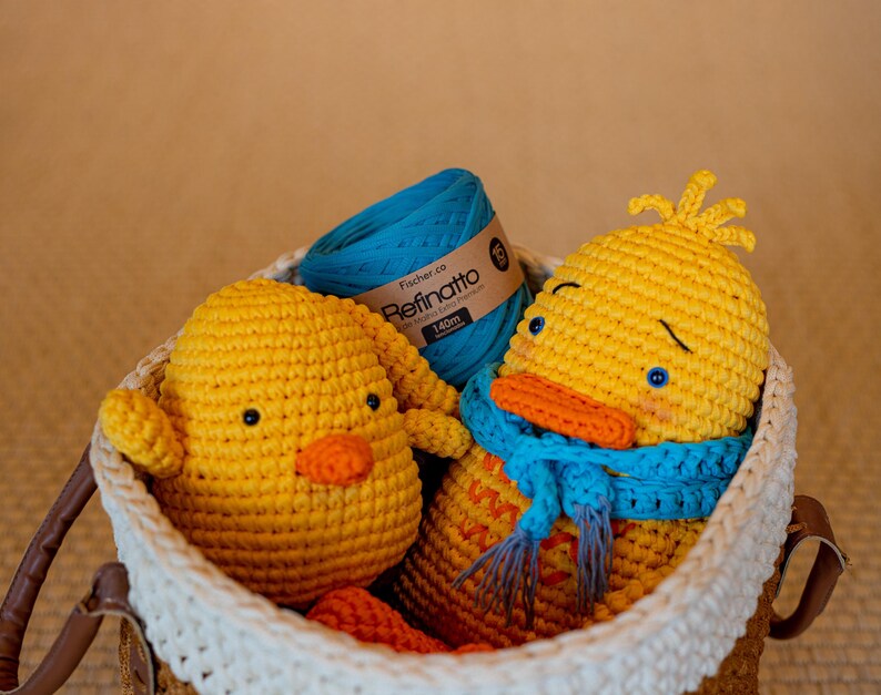 Peut inclure: Deux canetons au crochet jaunes, dont l'un porte une &eacute;charpe bleue, et une pelote de fil bleu sarcelle avec une &eacute;tiquette Fischer.co Refinatto 140m, le tout dans un panier tress&eacute; avec un bord blanc et des anses en similicuir marron.