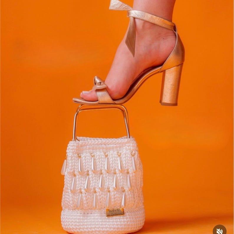 May include: A metallic gold block-heeled sandal with an ankle strap and a white crocheted bucket bag featuring a gold metal handle and dangling white teardrop-shaped beads. The bag has a small rectangular gold label on the front lower section.