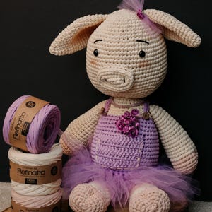 May include: A crocheted pig doll in a light beige color, wearing a purple overall dress with a tulle skirt and small purple flowers. Beside the doll are three stacked spools of Refinatto yarn in shades of purple, cream, and light pink.