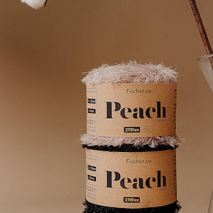 May include: Two spools of peach-colored yarn are stacked on a rock. The yarn is labeled "Peach" by Fischer.co, with "270Tex 100g" printed on the label. A cotton branch and a glass vase are in the background.