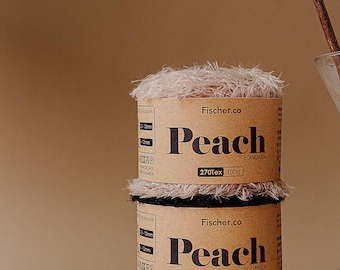 May include: Two spools of peach-colored yarn are stacked on a rock. The yarn is labeled "Peach" by Fischer.co, with "270Tex 100g" printed on the label. A cotton branch and a glass vase are in the background.