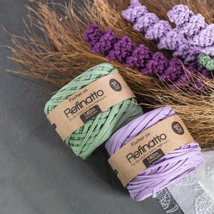 Peut inclure: Deux bobines de fil Fischer.co Refinatto en vert clair et violet clair, chacune entour&eacute;e d'une &eacute;tiquette en papier brun indiquant 15mm et 140m. Le fil est plac&eacute; pr&egrave;s d'herbes brunes s&eacute;ch&eacute;es et d'&eacute;l&eacute;ments d&eacute;coratifs en crochet violet.