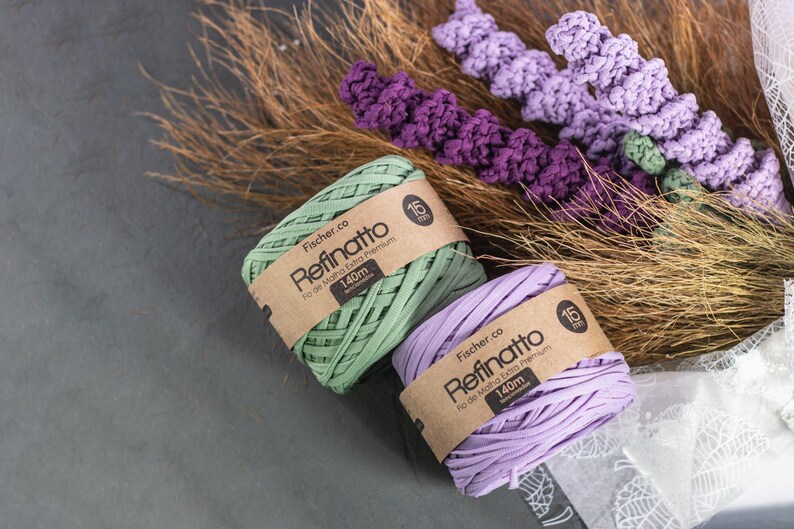 Peut inclure: Deux bobines de fil Fischer.co Refinatto en vert clair et violet clair, chacune entour&eacute;e d'une &eacute;tiquette en papier brun indiquant 15mm et 140m. Le fil est plac&eacute; pr&egrave;s d'herbes brunes s&eacute;ch&eacute;es et d'&eacute;l&eacute;ments d&eacute;coratifs en crochet violet.