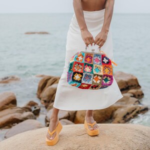 Peut inclure: Un sac &agrave; main dot&eacute; d'un motif de carr&eacute;s granny au crochet multicolores avec une fermeture &agrave; fermoir m&eacute;tallique argent&eacute;. Le sac est compos&eacute; de fils de diff&eacute;rentes couleurs, notamment rouge, bleu, jaune, vert et violet en motif carr&eacute;.