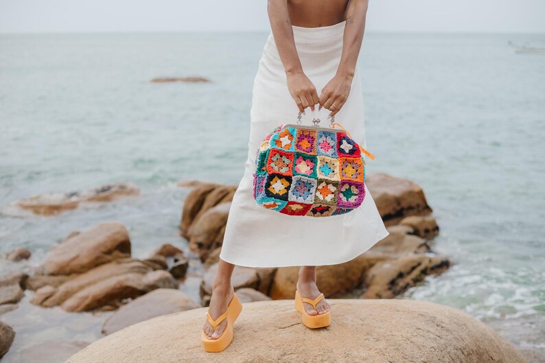 Peut inclure: Un sac &agrave; main dot&eacute; d'un motif de carr&eacute;s granny au crochet multicolores avec une fermeture &agrave; fermoir m&eacute;tallique argent&eacute;. Le sac est compos&eacute; de fils de diff&eacute;rentes couleurs, notamment rouge, bleu, jaune, vert et violet en motif carr&eacute;.