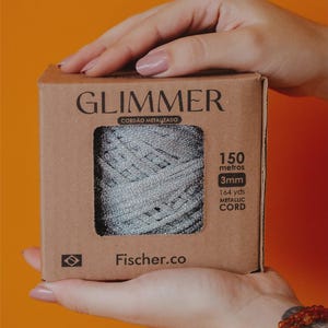 May include: A box of metallic cord held by hands. The box is brown with the word "GLIMMER" printed on the front. The cord is silver and measures 3mm in diameter and 164 yards in length.