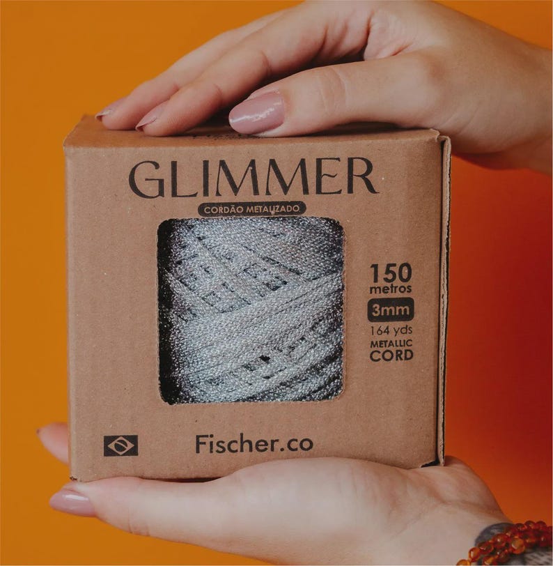 May include: A box of metallic cord held by hands. The box is brown with the word "GLIMMER" printed on the front. The cord is silver and measures 3mm in diameter and 164 yards in length.