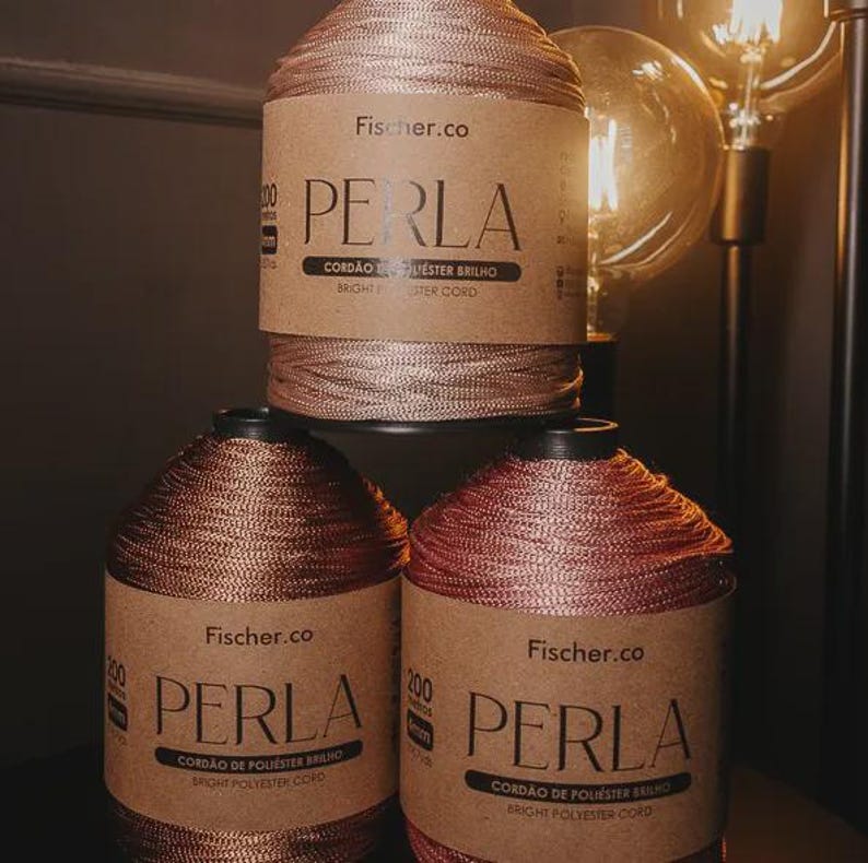May include: Three spools of Fischer.co Perla bright polyester cord stacked in a pyramid. Each spool features a tan paper label with the text PERLA, CORD&Atilde;O DE POLI&Eacute;STER BRILHO, and BRIGHT POLYESTER CORD. The cord has a metallic, shimmering finish.