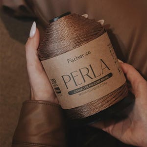 May include: A spool of metallic bronze polyester cord labeled Fischer.co PERLA Bright Polyester Cord. The label indicates 200 meters and 4mm thickness. The cord has a shiny, smooth texture wrapped tightly around a black cylindrical core.