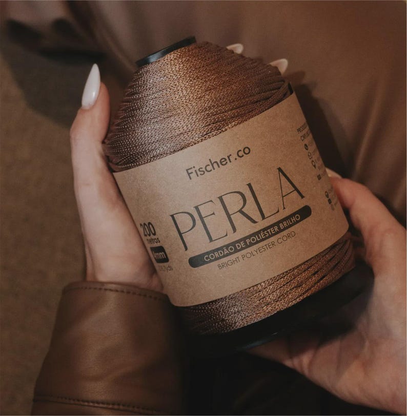 May include: A spool of metallic bronze polyester cord labeled Fischer.co PERLA Bright Polyester Cord. The label indicates 200 meters and 4mm thickness. The cord has a shiny, smooth texture wrapped tightly around a black cylindrical core.