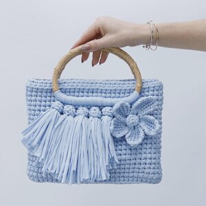 Peut inclure: Un sac &agrave; main rectangulaire en crochet bleu clair avec un motif en relief, une anse ronde en bambou, une rang&eacute;e de pompons en tissu et une grande fleur en crochet fix&eacute;e sur le devant du sac.