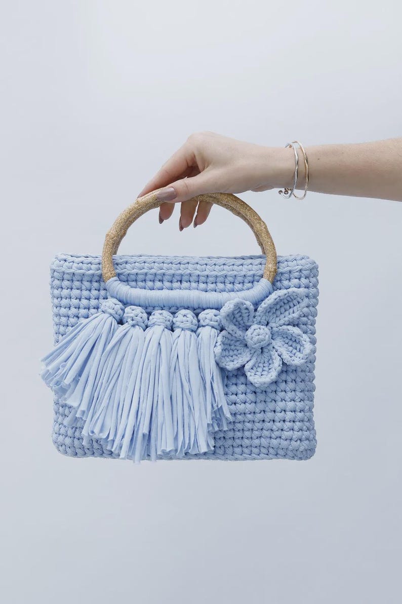 Peut inclure: Un sac &agrave; main rectangulaire en crochet bleu clair avec un motif en relief, une anse ronde en bambou, une rang&eacute;e de pompons en tissu et une grande fleur en crochet fix&eacute;e sur le devant du sac.